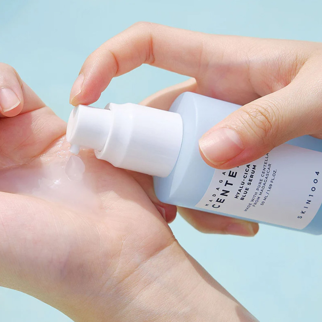Hand applying skincare serum from a bottle labeled 'SKIN1004' on a light blue background