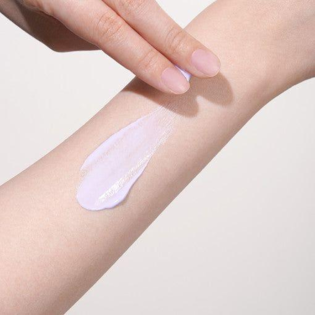 Hand applying a light purple cream or lotion to an arm on a plain background