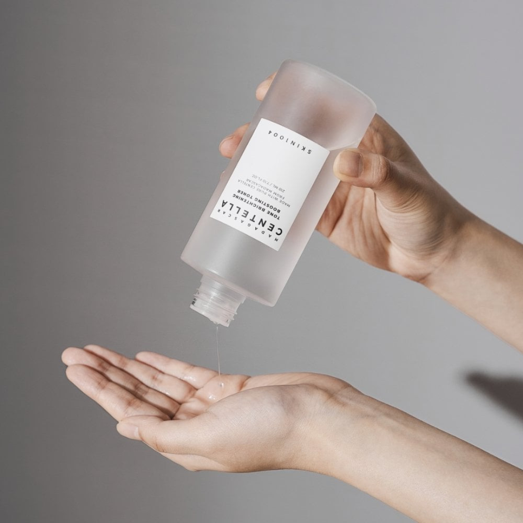 Hand holding a clear bottle with a label, dispensing liquid onto another hand against a gray background