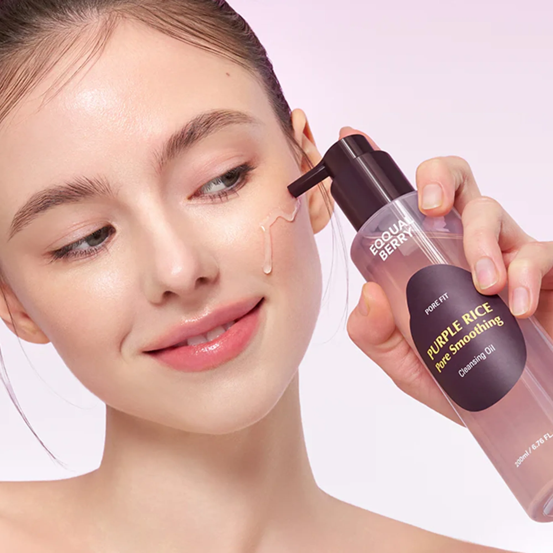 Woman applying cleansing oil to her face with a bottle labeled 'Purple Rice Pore Smoothing' on a light pink background.