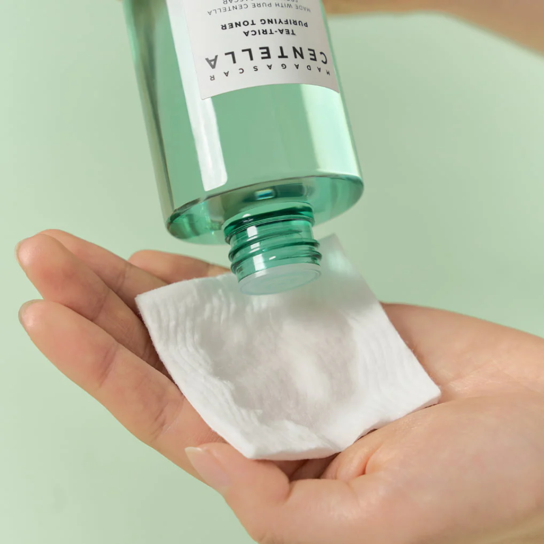 Hand holding a bottle of Centella Asiatica toner over a cotton pad with a light green background.