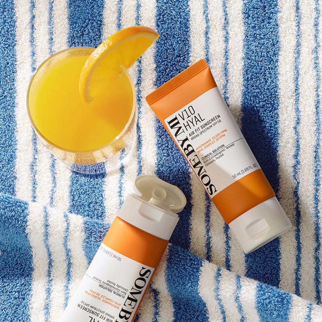Two tubes of 'SOMEBYMI' sunscreen on a blue and white striped towel with a glass of orange juice.