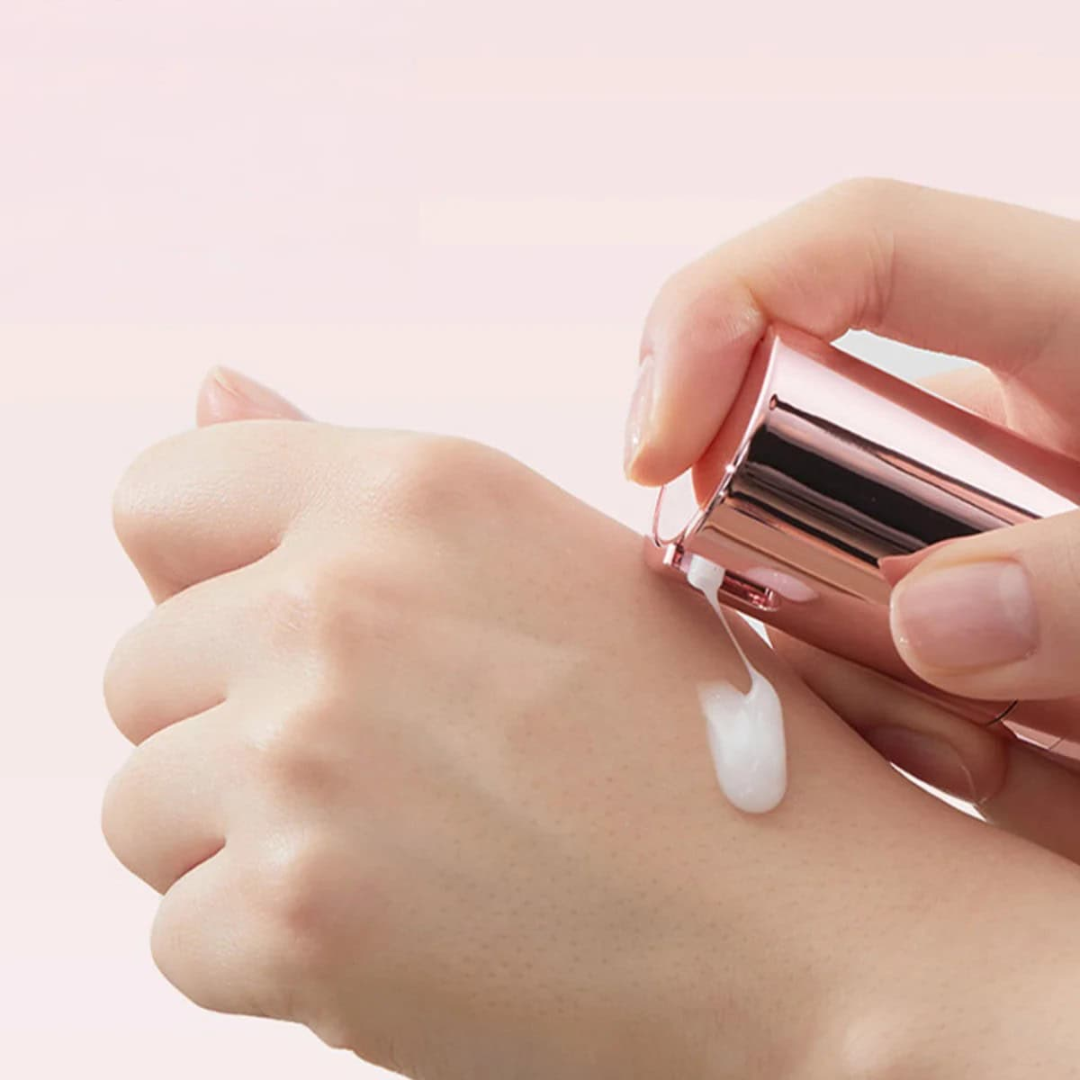 Hand applying cream from a pink tube onto a wrist against a light pink background